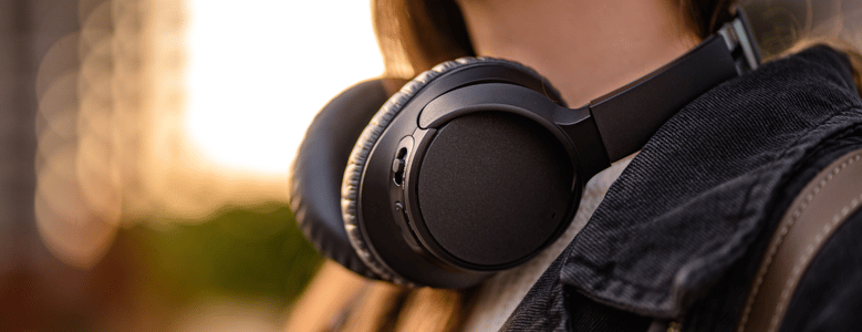 Detalhe de um fone de ouvido over-ear preto pendurado no pescoço de uma pessoa que usa jaqueta jeans escura. Ao fundo, ambiente externo desfocado com luz dourada do fim do dia.