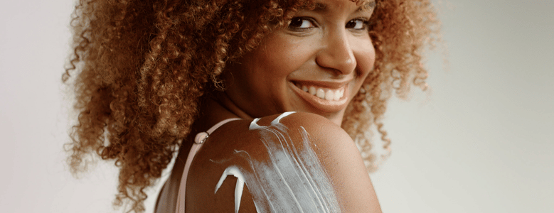 Mulher negra de cabelos cacheados sorrindo enquanto aplica hidratante corporal no ombro.