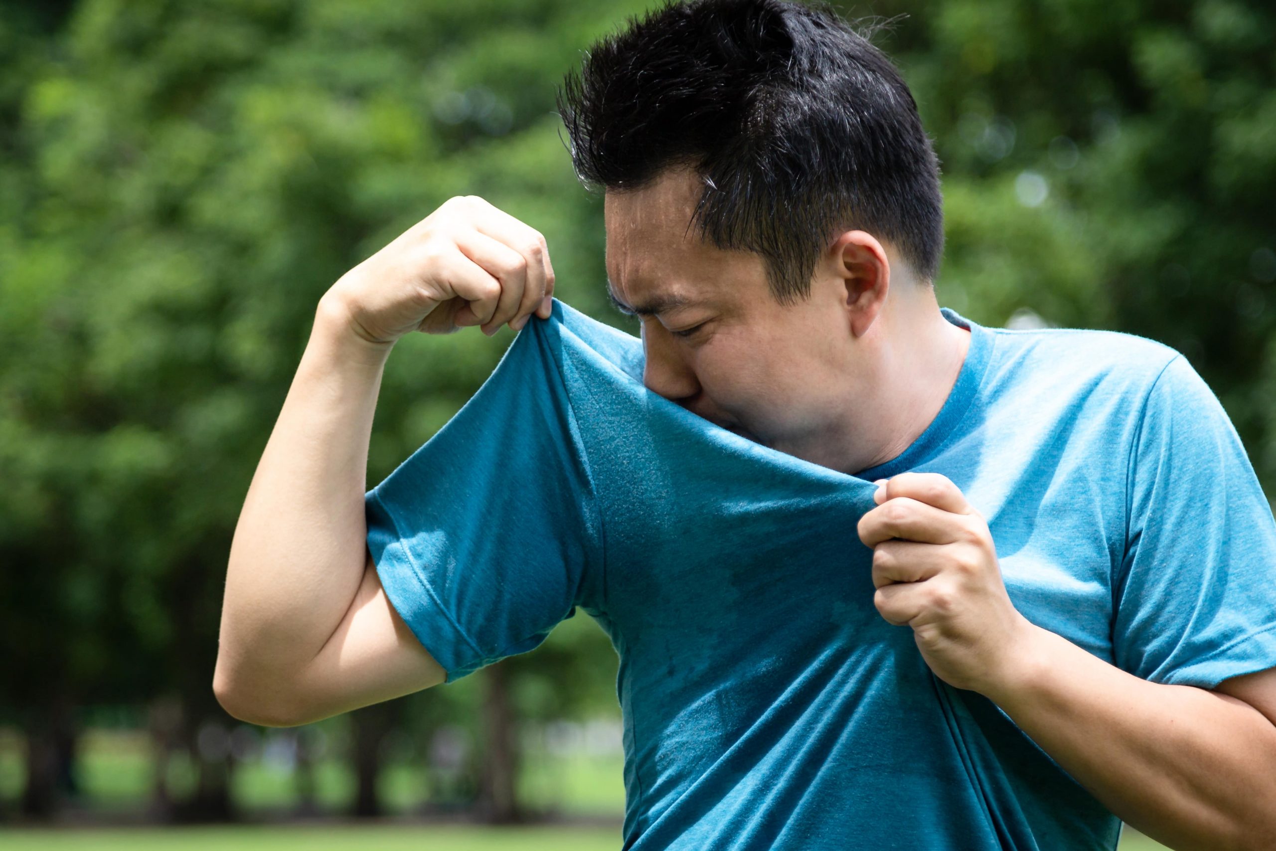 Homem com o rosto virado para a esquerda, vestido com uma camiseta esportiva azul molhada de suor. Com as mãos ele aproxima o tecido do rosto e faz expressão de desagrado.